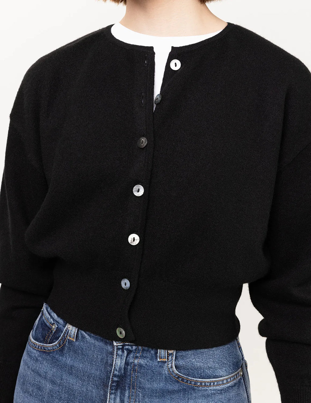 Black cardigan with white buttons worn over a white shirt, paired with blue jeans on a plain background. - Black