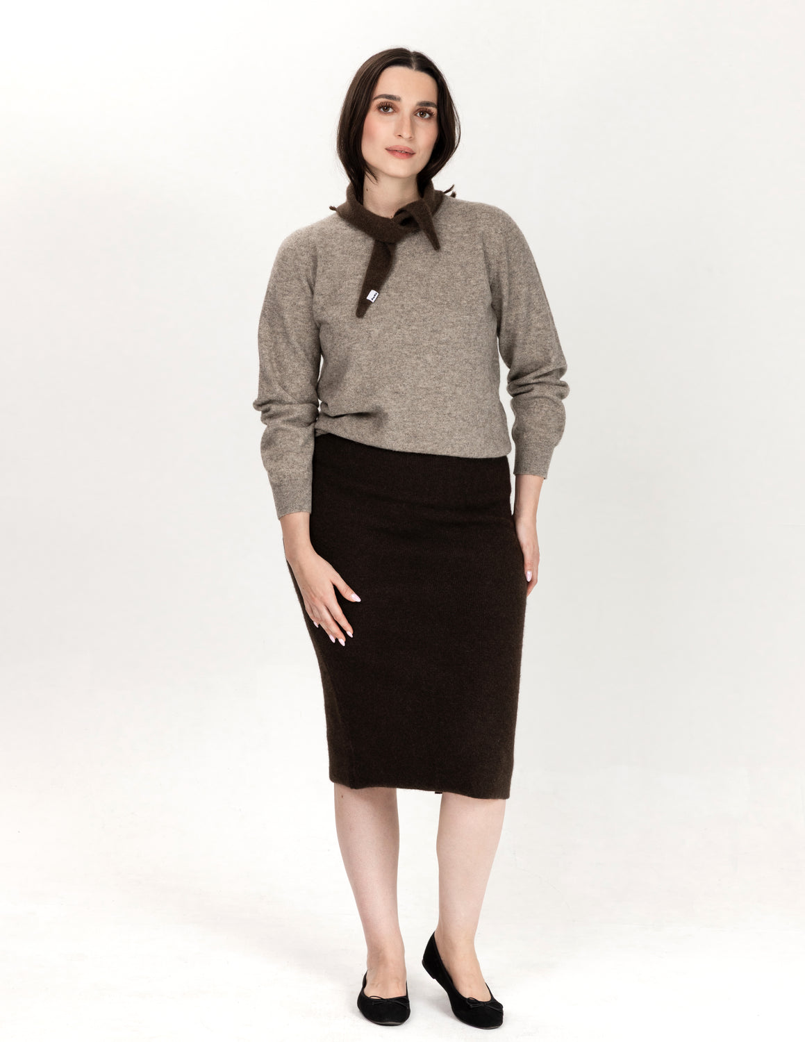 A woman modeling a gray knit sweater with a dark brown necktie detail, paired with a dark brown knee-length skirt and black flats, against a plain white studio background.