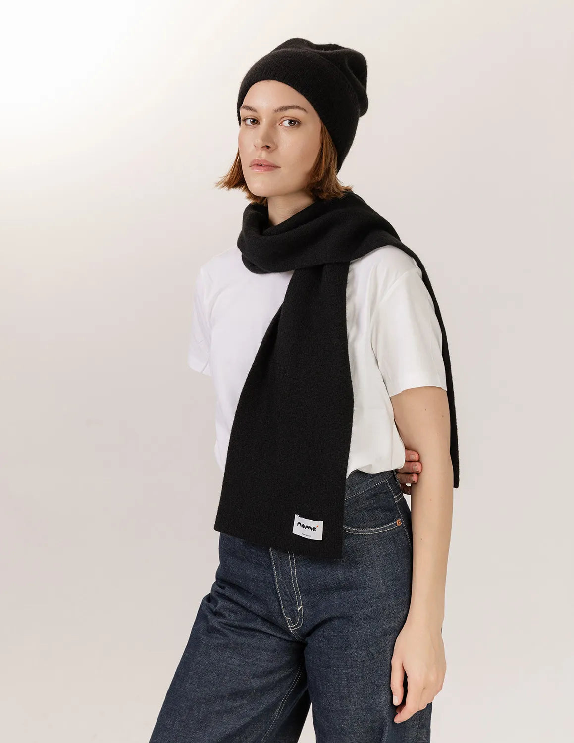 Person wearing a black scarf, white shirt, and dark jeans on a plain background -Black