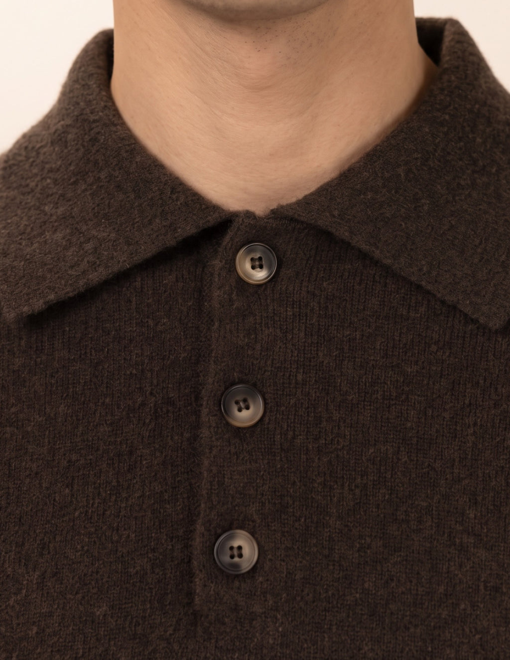 Close-up of a person wearing a dark brown knit polo shirt with a collar and three dark buttons, shown against a plain light beige background - Natural Brown