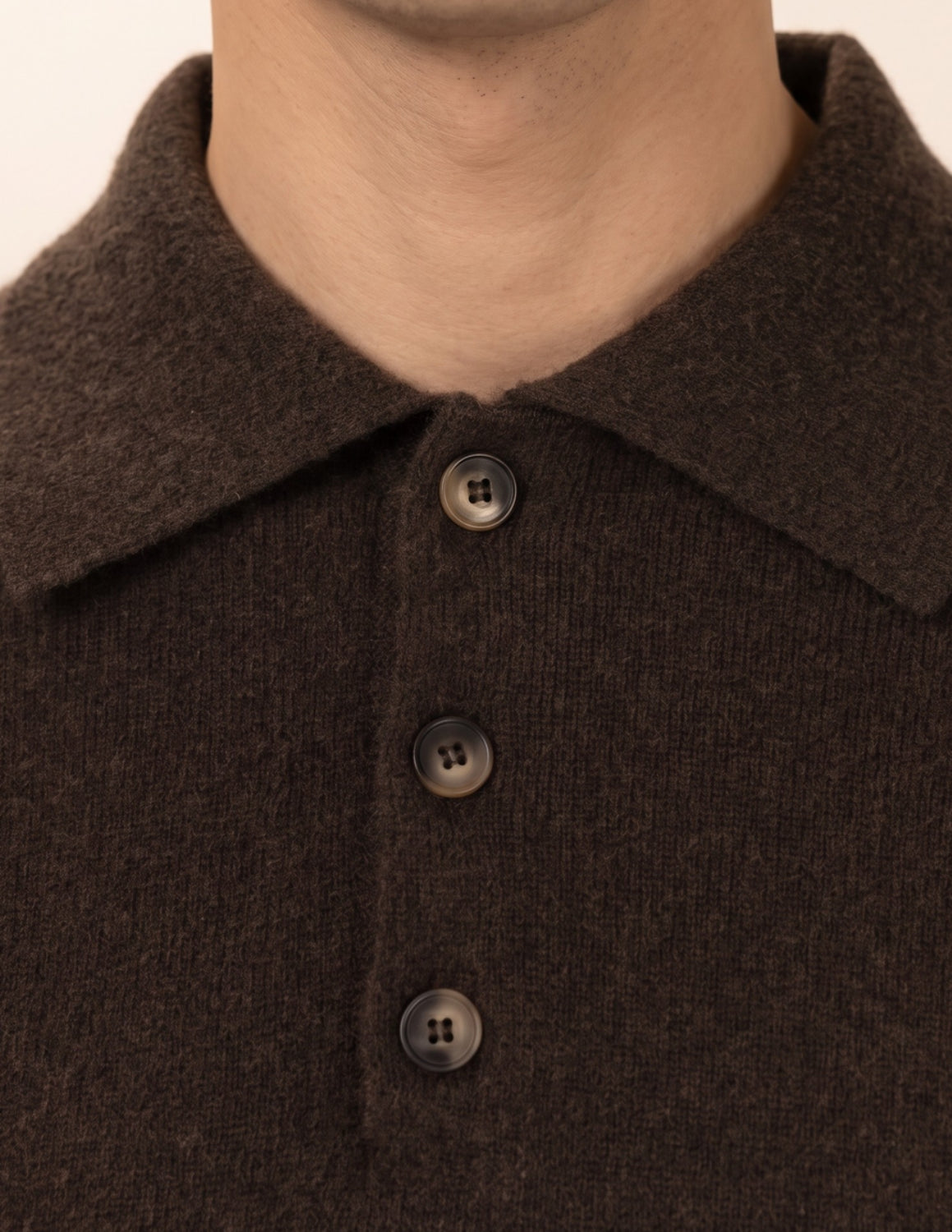 Close-up of a person wearing a dark brown knit polo shirt with a collar and three dark buttons, shown against a plain light beige background - Natural Brown