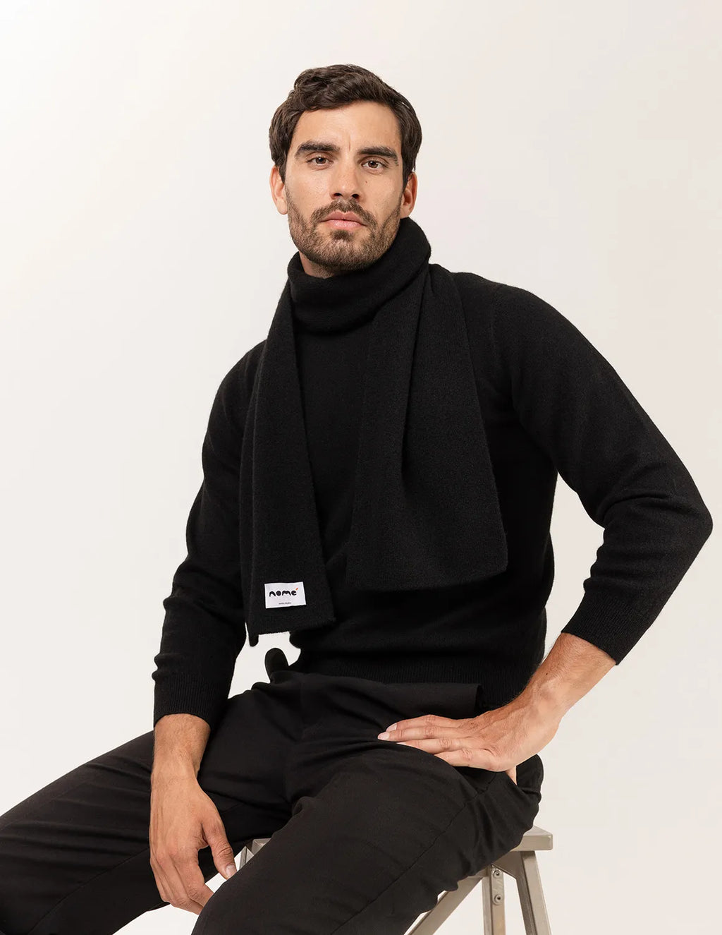 Man wearing a black outfit with a visible brand label, sitting on a stool against a plain background - Black